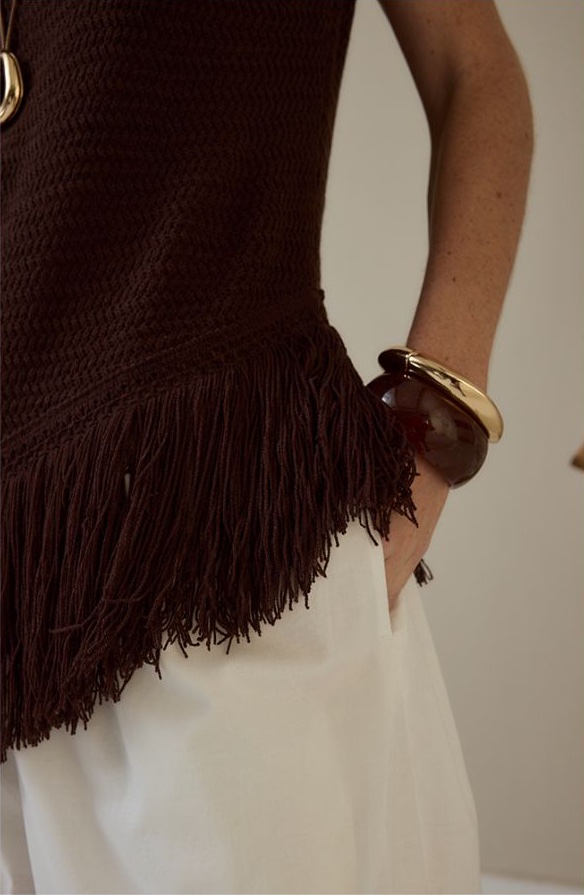 Close-up of a brown fringed crochet top with gold and tortoiseshell bangles over white trousers