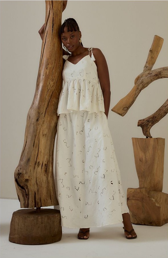 Model in a white broderie camisole and maxi skirt leaning against a driftwood sculpture